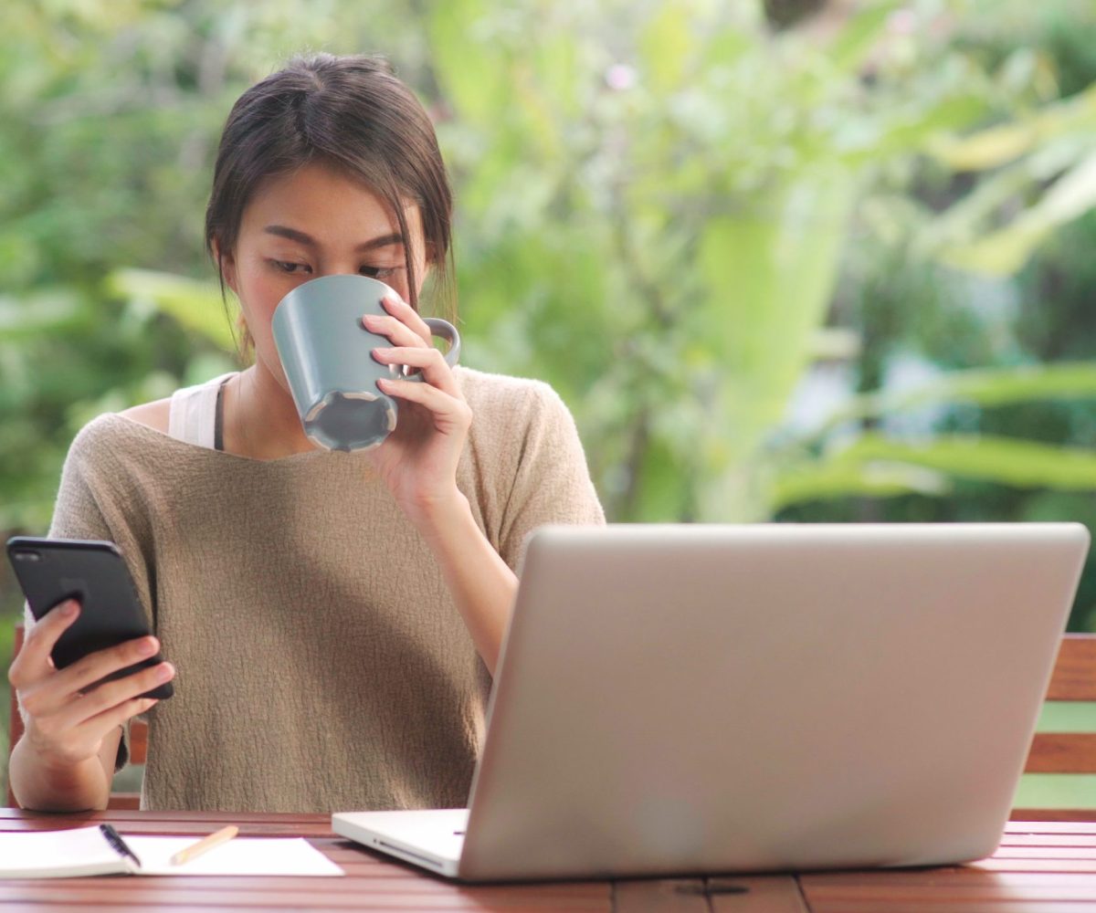 Freelance Asian woman working at home, business female working on laptop and using mobile phone drinking coffee sitting on table in the garden in morning. Lifestyle women working at home concept.